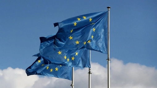 Two European Union flags with yellow stars flying on flagpoles against a bright blue sky with clouds