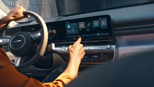 Driver's hand touching a wide in-car touchscreen displaying app icons and a digital instrument cluster