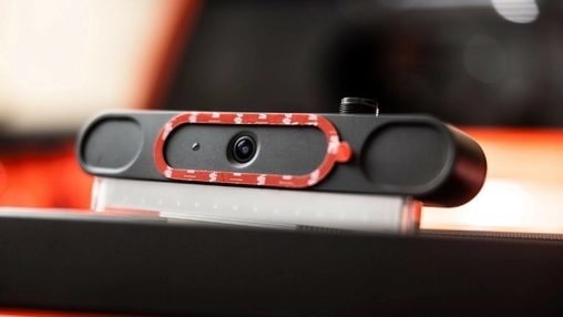 Black webcam module with lens ringed in red tape, mounted on a laptop edge against blurred red lights